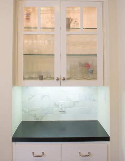 A built-in kitchen cabinet with frosted glass doors above a black countertop with white drawers and a backsplash, illuminated by under-cabinet lighting.