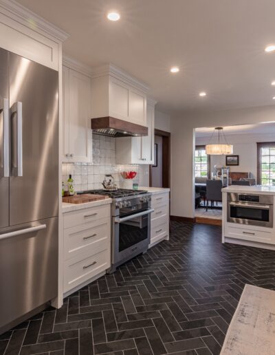 Modern kitchen with stainless steel appliances, white cabinets, and dark herringbone floor tiles, leading to an adjoining living area.