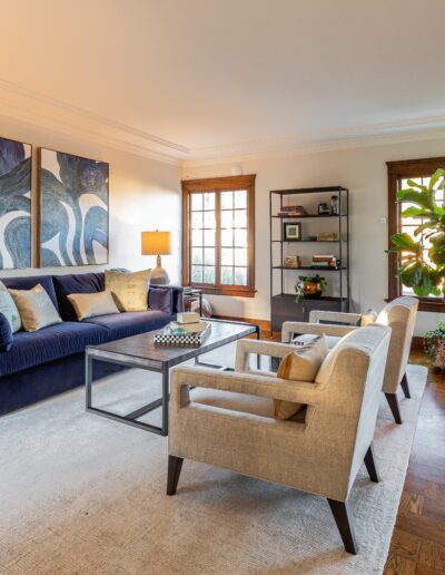 Elegant living room with a blue sofa, two beige armchairs, a large wall painting, and several plants, featuring hardwood floors and natural lighting.