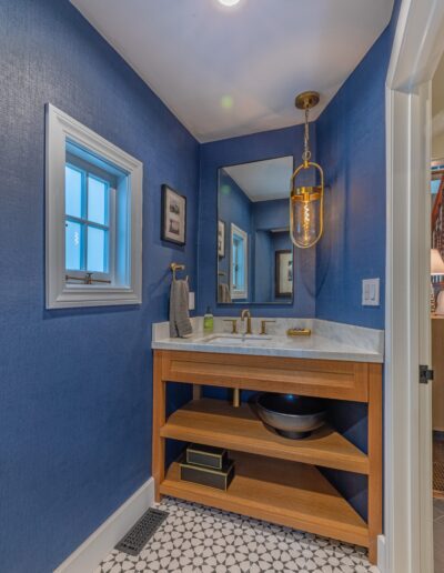 A modern bathroom featuring deep blue walls, geometric floor tiles, and a wooden vanity with a circular basin, illuminated by a hanging light fixture.