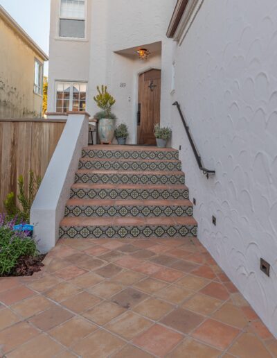 Front entrance of a house with decorative tiled steps leading to a wooden door, flanked by potted plants, under a clear sky.