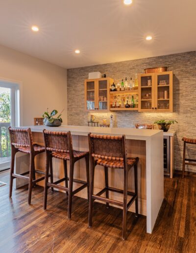 Modern home bar area with stone wall, wooden flooring, a white bar counter with four wicker bar stools, and a view of trees through open sliding doors.