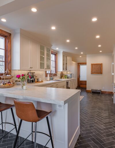 Modern kitchen interior with white cabinetry, marble island, bar stools, and dark herringbone floor. brightly lit with natural light coming from wood-framed windows.